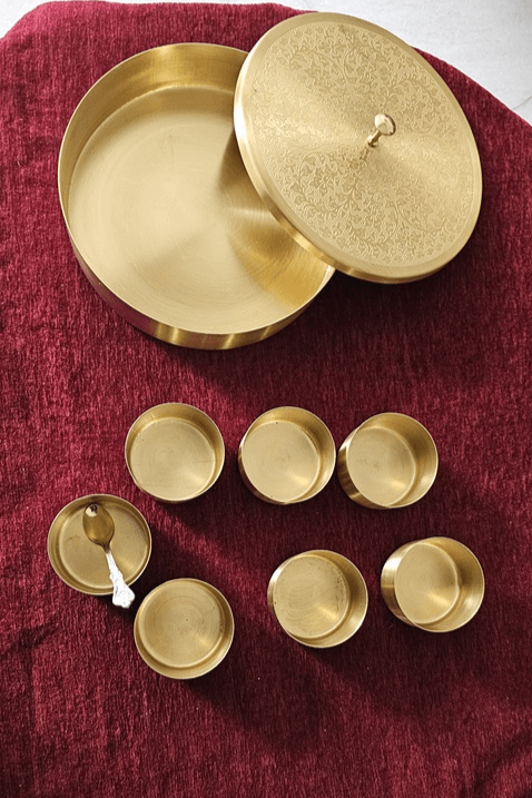 Complete brass masala dabba set including etched lid, seven small containers, and a spoon, placed on red fabric – premium spice storage by Troika.