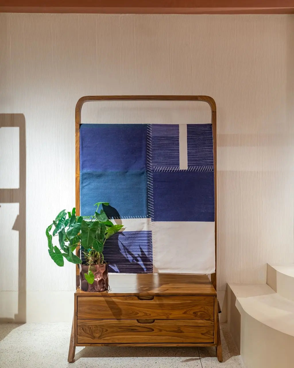 Wooden standing unit with blue and white patchwork dhurrie display and potted plant, placed against neutral backdrop – home decor by Troika.