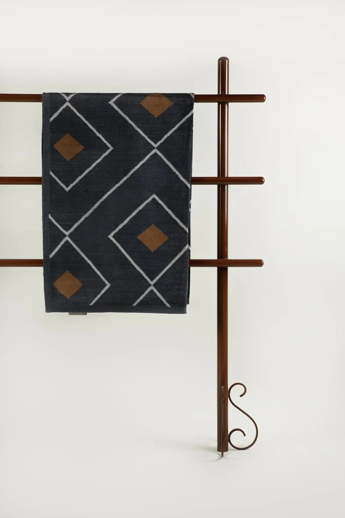 Wide view of the dark grey dhurrie on a wooden rack with an ornate base, placed against a plain white wall.