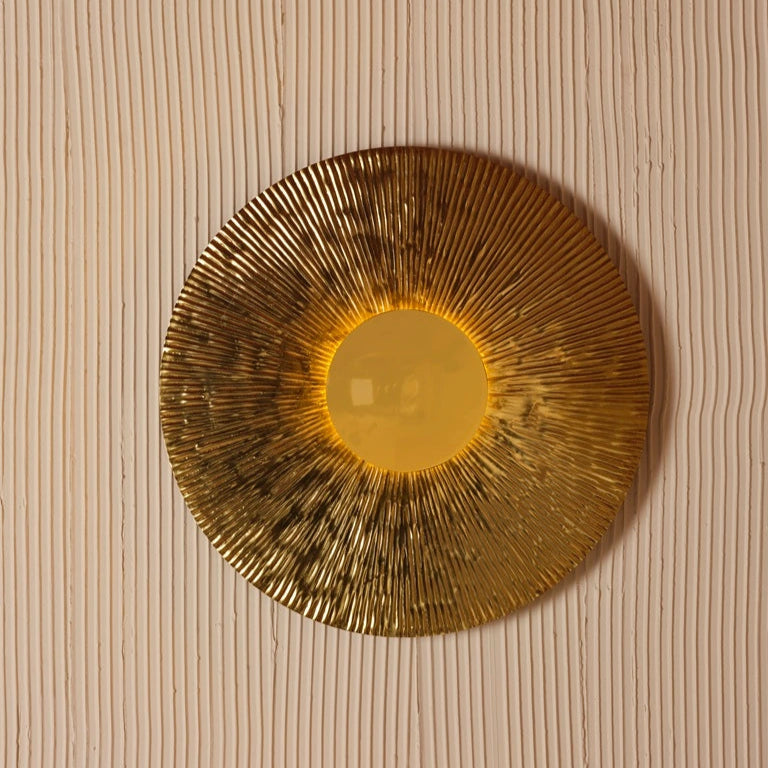 Round gold wall light with radial texture and central diffuser, mounted on ribbed beige wall – radiant decorative lighting by Troika.