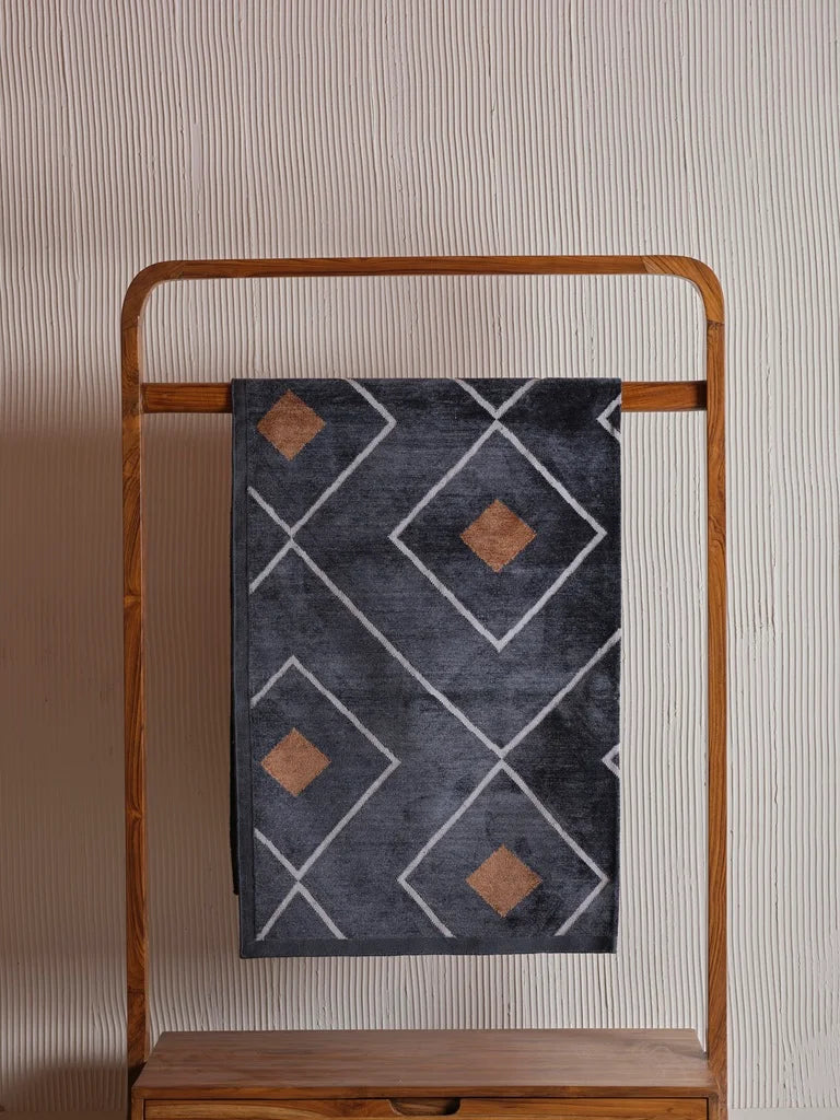 Dark grey durrie rug with geometric white and copper patterns, draped on a wooden rack. Modern and earthy accent for any room.