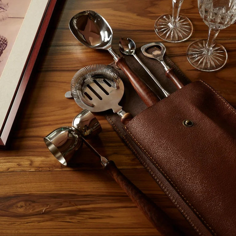 Luxury bar tool set with wooden handles in leather case, styled on walnut wood surface with glassware.