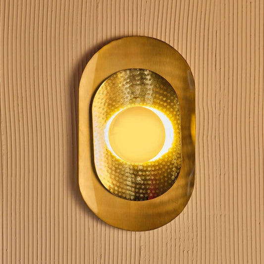 Oval-shaped gold wall light with hammered brass texture and diffused center glow, mounted on vertical ribbed wall – luxury lighting fixture by Troika.