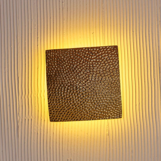 Gold square wall light fixture glowing warmly handcrafted tamper-textured metal lighting piece.